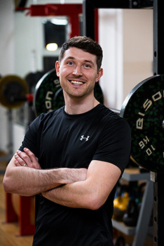 Jonny Harper, a young white man stands in front of a weight rack in a gym.