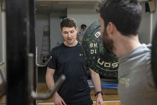 A man lifts a weight in the foreground while a physiotherapist watches.